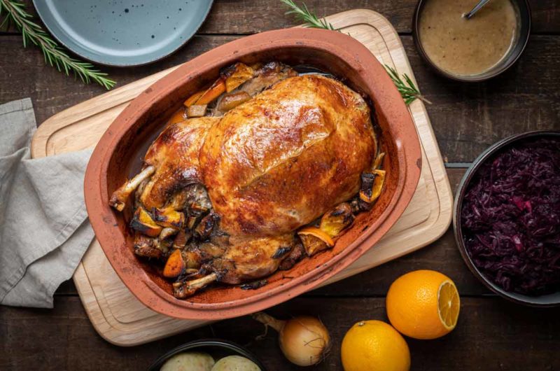Gebratene Ente im Bräter, serviert mit Rotkohl, Klößen und Orangen auf einem Holztisch mit Teller und Soße.