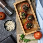 Gebackene gefüllte Tomaten in einer Holzform, umgeben von Zwiebel, Pfeffer, Kirschtomaten und Petersilie auf einem Tisch.