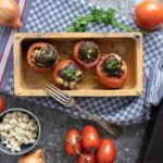 Gefüllte Tomaten in einer Form auf Küchentuch, umgeben von Tomaten, Zwiebeln und Schafskäsewürfeln.