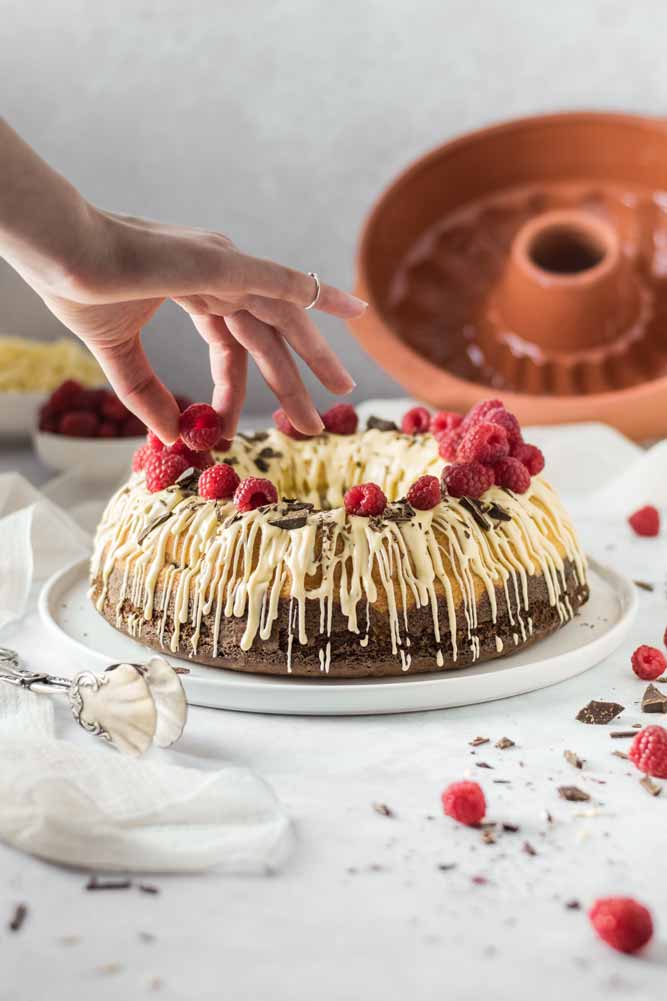 Hand dekoriert Kranzkuchen mit Himbeeren und weißer Schokolade; Gugelhupfform im Hintergrund.