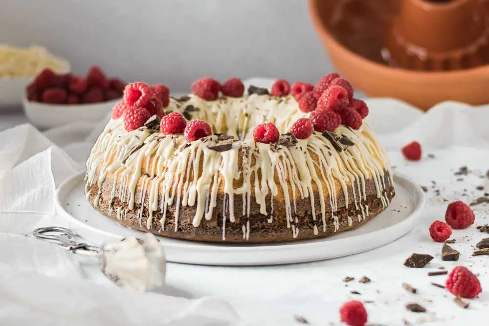 Gugelhupf mit weißer Schokolade, Himbeeren und Schokoladenstücken garniert auf einem Teller.