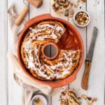 Nusszopf in einer roten Form auf weißem Holztisch, dekoriert mit Nüssen und Zuckerguss, mit Messer und Backutensilien.