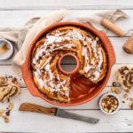 Runder Hefezopf mit Zuckerguss und Nüssen in einer Backform, daneben ein Holzroller und ein angeschnittenes Stück.