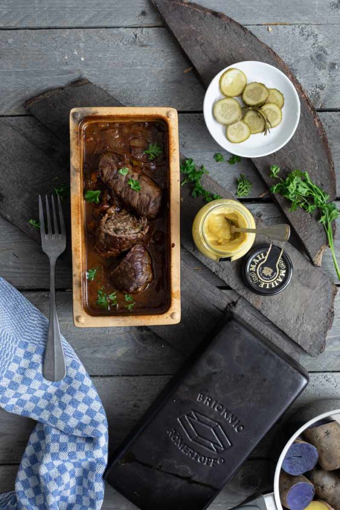 Rinderrouladen in einer Backform mit Soße, eingelegte Gurken, Senfglas, und Kräuter auf einem Holztisch.