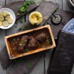 Rinderbraten in einer Backform mit Essiggurken, Senf und Kartoffeln auf einem Holztisch arrangiert.