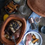 Ton-Topf mit Braten, Süßkartoffelwedges, Gewürzen und Pfeffermühle auf Holztisch. Teller mit Fleisch und Beilage.
