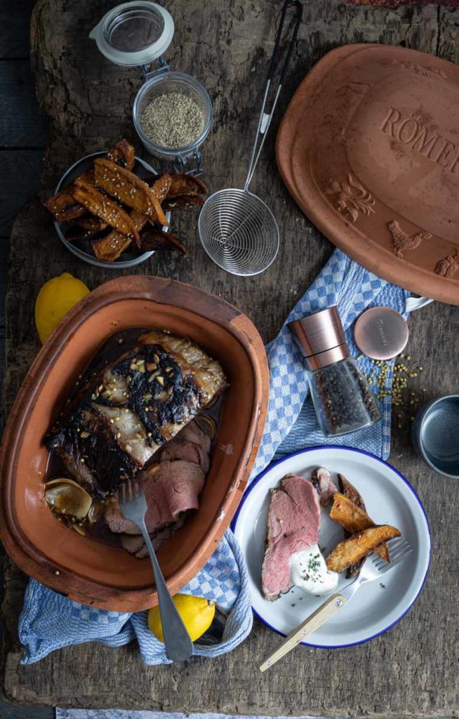 Ton-Topf mit Braten, Süßkartoffelwedges, Gewürzen und Pfeffermühle auf Holztisch. Teller mit Fleisch und Beilage.