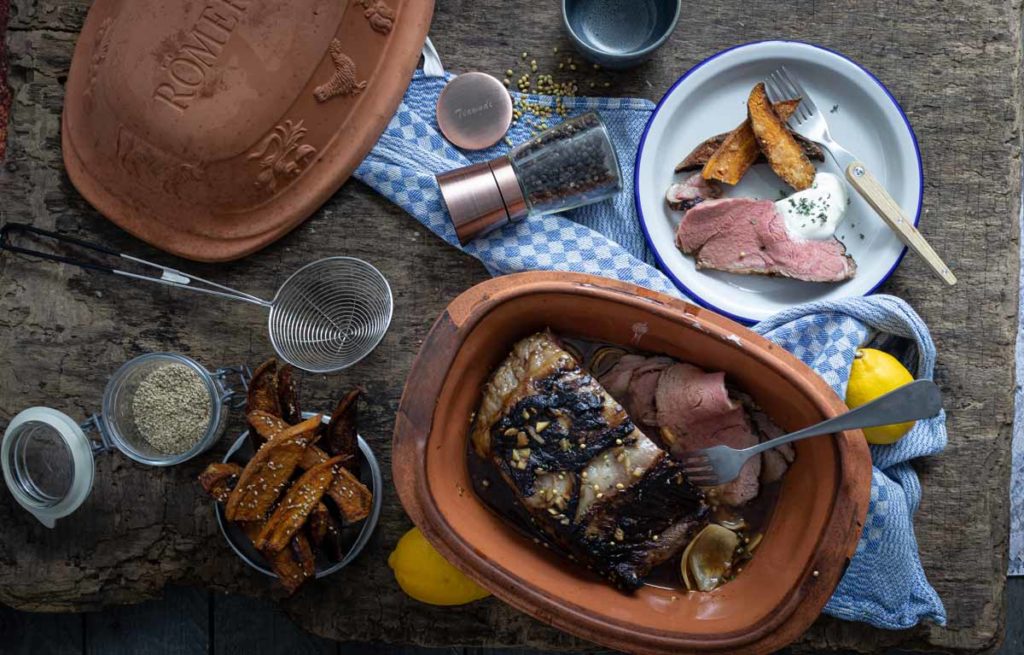 Bräter mit Braten, serviert mit Kartoffelspalten und Sauce, auf Holzbrett mit Gewürzen und Küchenutensilien arrangiert.