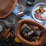 Bräter mit Braten, serviert mit Kartoffelspalten und Sauce, auf Holzbrett mit Gewürzen und Küchenutensilien arrangiert.