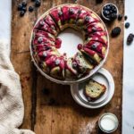 Kuchenkranz mit Beerenglasur auf Holzbrett, umgeben von frischen Beeren, mit einem Glas Milch und Puderzucker.