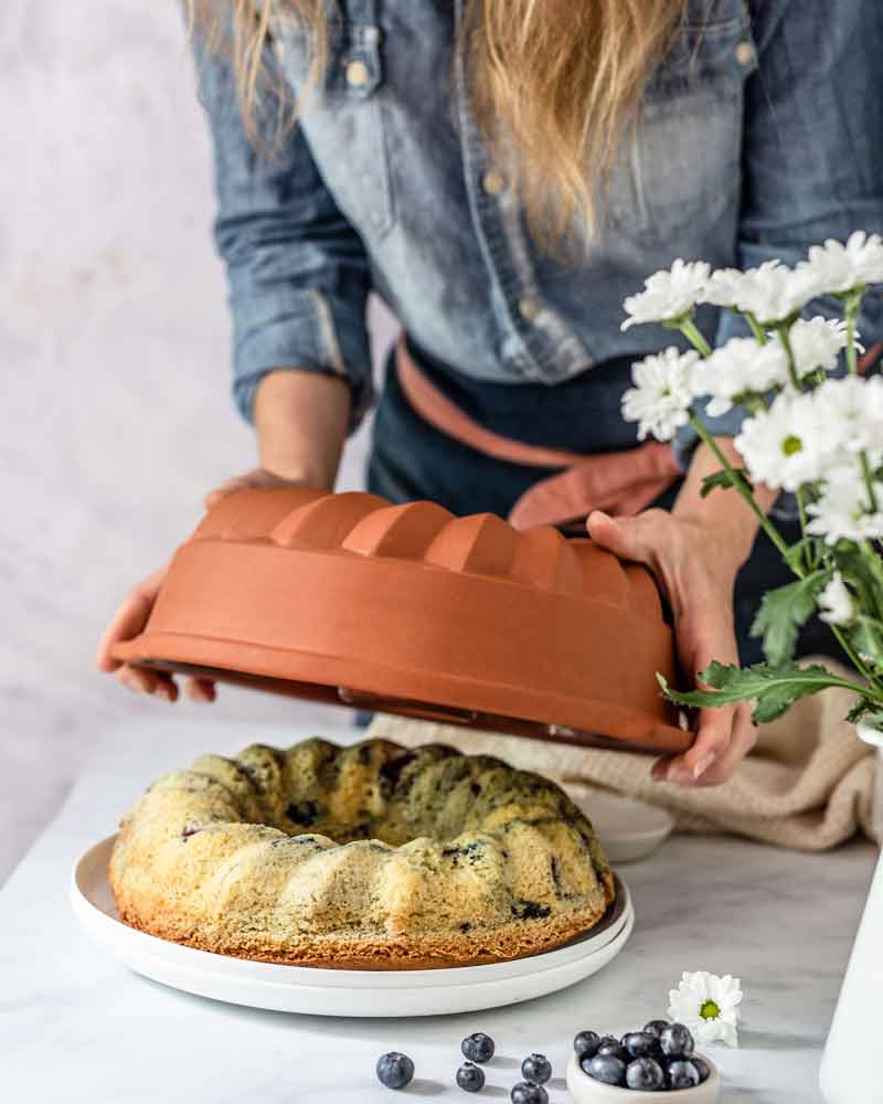 Person deckt frisch gebackenen Gugelhupf auf einem Teller ab, daneben Blumen und Blaubeeren.