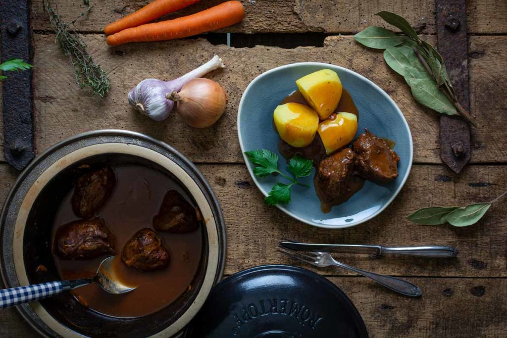 Eintopf mit Fleisch in Soße, serviert mit Kartoffeln auf blauem Teller. Zutaten wie Karotten und Knoblauch daneben.