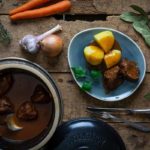 Eintopf mit Fleisch in Soße, serviert mit Kartoffeln auf blauem Teller. Zutaten wie Karotten und Knoblauch daneben.