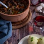Eintopf im Keramiktopf mit Kartoffelknödeln auf Teller und Rotwein im Glas auf Holztisch.
