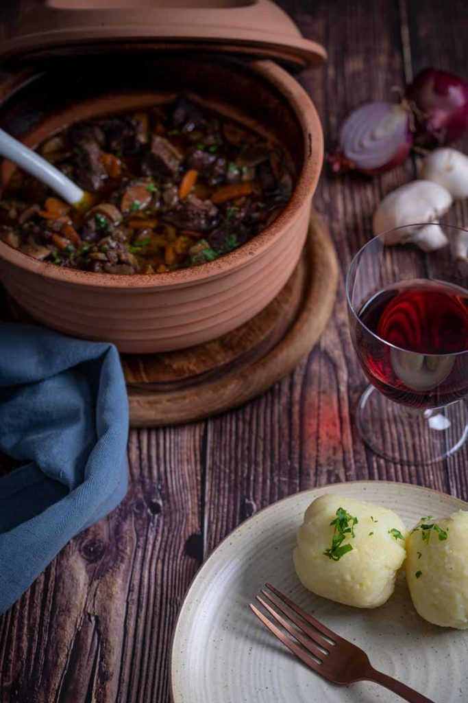 Eintopf im Keramiktopf mit Kartoffelknödeln auf Teller und Rotwein im Glas auf Holztisch.