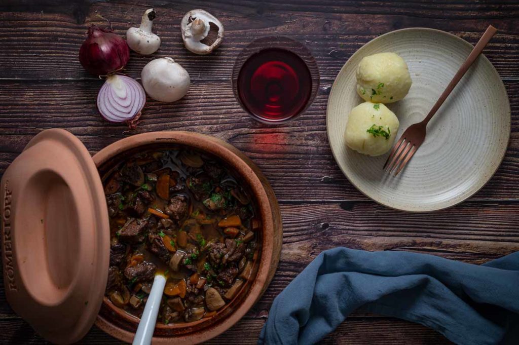 Tonkrug mit Rindereintopf, zwei Kartoffelknödel auf Teller, Rotwein, Zwiebeln und Champignons auf Holzuntergrund.