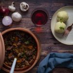 Tonkrug mit Rindereintopf, zwei Kartoffelknödel auf Teller, Rotwein, Zwiebeln und Champignons auf Holzuntergrund.