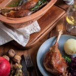 Gebratene Gänsekeule mit Knödel, Rotkohl und Rosmarin auf Teller. Daneben Apfel, Nüsse und Bräter mit Gans.