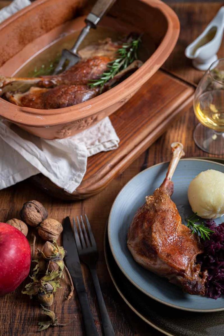 Gebratene Gänsekeule mit Knödel, Rotkohl und Rosmarin auf Teller. Daneben Apfel, Nüsse und Bräter mit Gans.