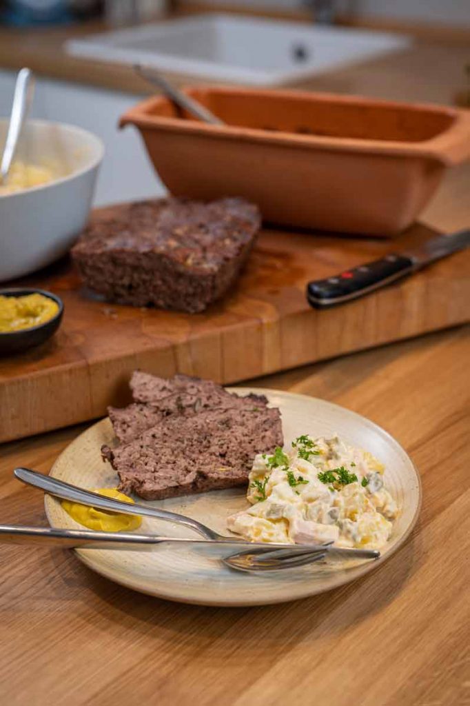 Leberkäse und Kartoffelsalat auf einem Teller, mit Senf und Besteck, auf einem Holztisch angerichtet.