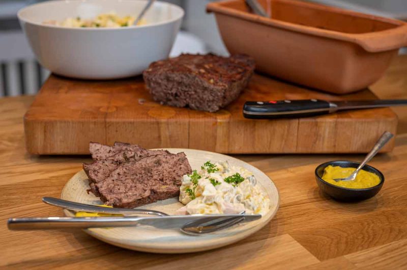 Scheiben Hackbraten mit Kartoffelsalat auf einem Teller, daneben Senf, auf Holztisch mit Schneidebrett im Hintergrund.