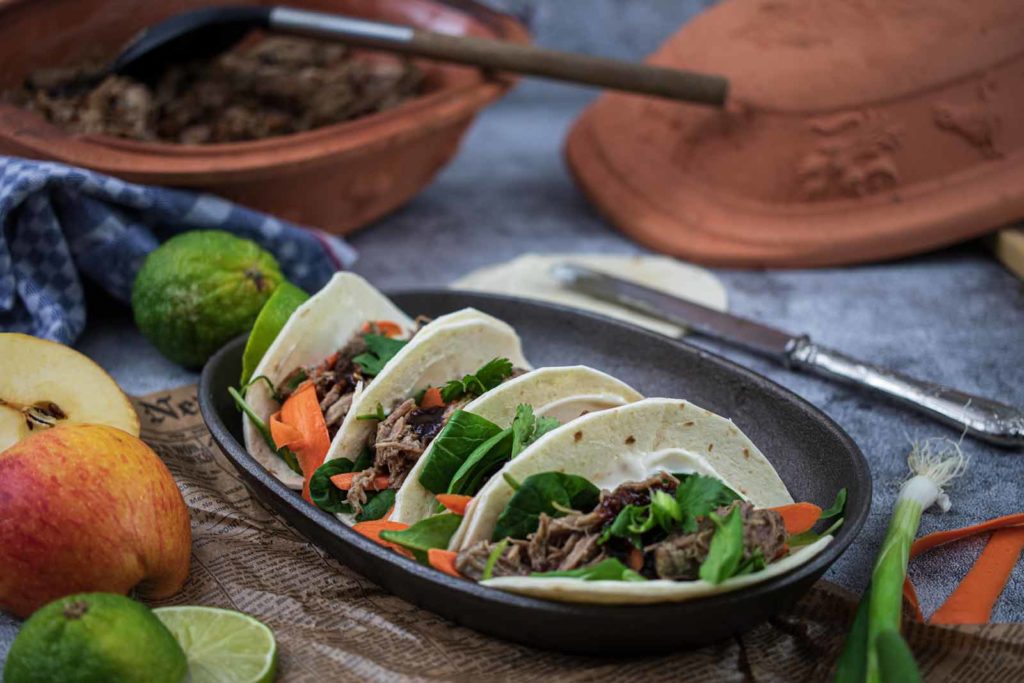 Drei Tacos mit Rindfleisch, Karotten und Salat in einer Schale, umgeben von Limetten und einem Apfel.