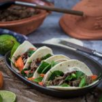 Drei Tacos mit Rindfleisch, Karotten und Salat in einer Schale, umgeben von Limetten und einem Apfel.