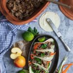 Schmorgericht im Tontopf, angerichtete Tacos mit Limette und Apfel, auf grauem Untergrund mit kariertem Handtuch.
