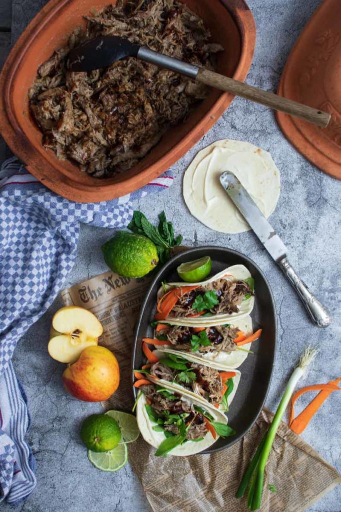 Schmorgericht im Tontopf, angerichtete Tacos mit Limette und Apfel, auf grauem Untergrund mit kariertem Handtuch.