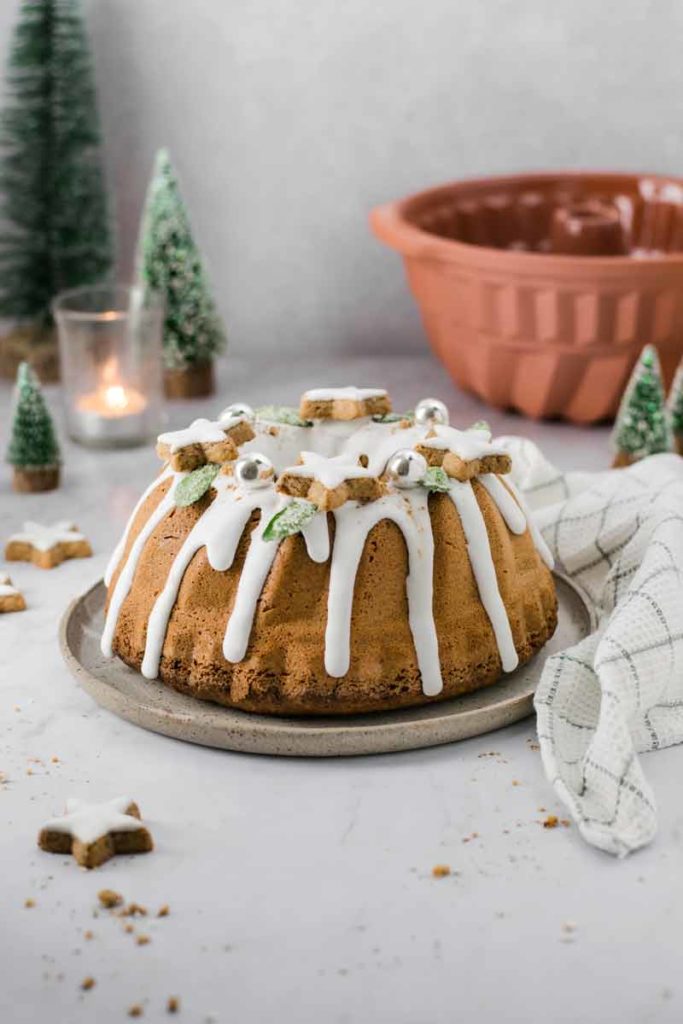 Weihnachtlicher Gugelhupf mit Zuckerguss, Plätzchen und Winterdeko auf einem Tisch mit Kerze und Tannenbäumchen.