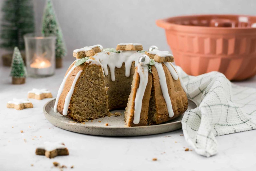 Ein Gugelhupf mit Zuckerguss und Keksen dekoriert, auf einem Teller, im Hintergrund ein Tuch und Kerzenlicht.