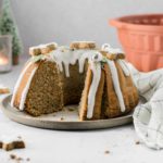 Ein Gugelhupf mit Zuckerguss und Keksen dekoriert, auf einem Teller, im Hintergrund ein Tuch und Kerzenlicht.