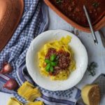 Teller mit Tagliatelle und Bolognese, umgeben von Rosmarin, Parmesan und Schalotten auf kariertem Tuch.