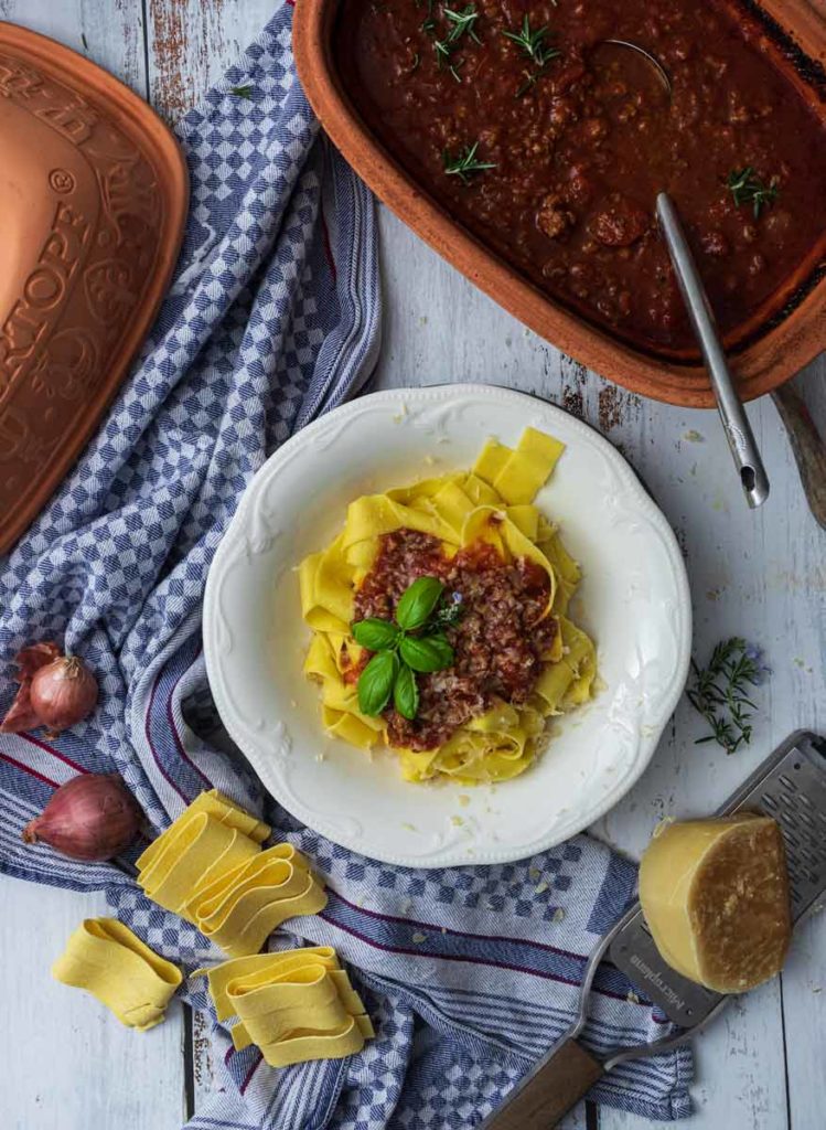 Teller mit Tagliatelle und Bolognese, umgeben von Rosmarin, Parmesan und Schalotten auf kariertem Tuch.