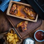 Ofengebackene Hähnchenflügel auf Holzbrett, mit Pommes, Mayonnaise und Ketchup, daneben ein blau kariertes Tuch.