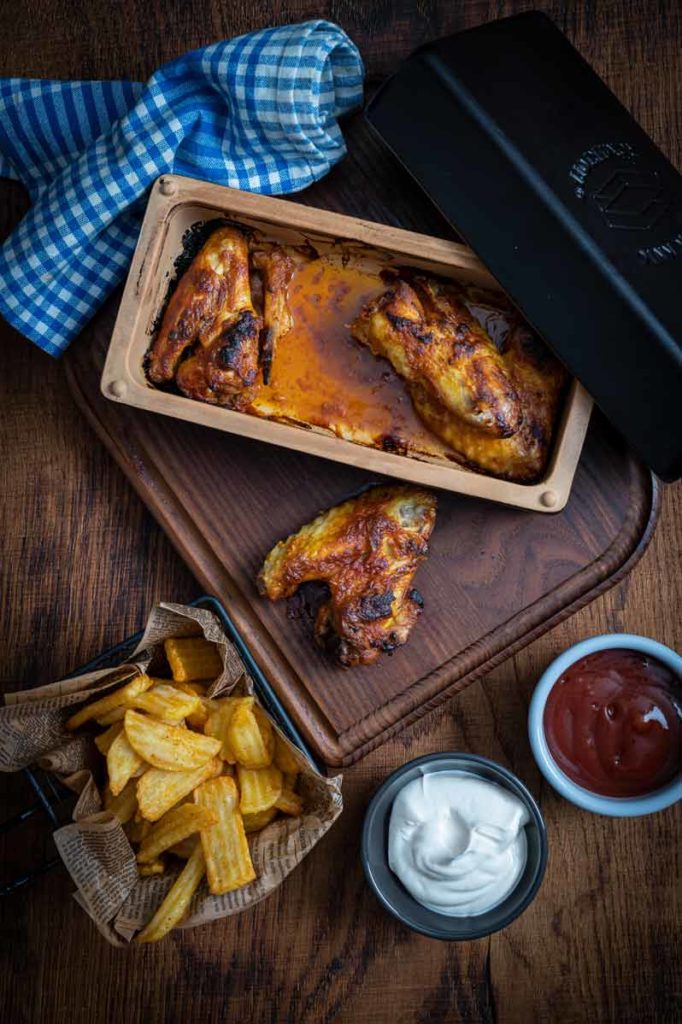 Ofengebackene Hähnchenflügel auf Holzbrett, mit Pommes, Mayonnaise und Ketchup, daneben ein blau kariertes Tuch.