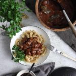 Teller mit Nudeln und Fleischragout, daneben Topf und Schüssel mit dunkler Sauce, Gabel, Petersilie, graues Tuch.