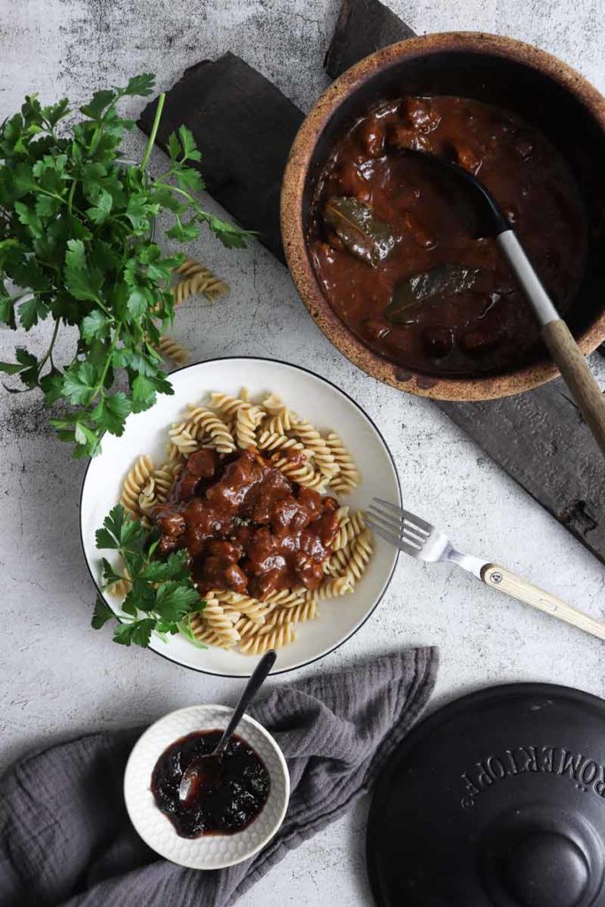 Teller mit Nudeln und Fleischragout, daneben Topf und Schüssel mit dunkler Sauce, Gabel, Petersilie, graues Tuch.