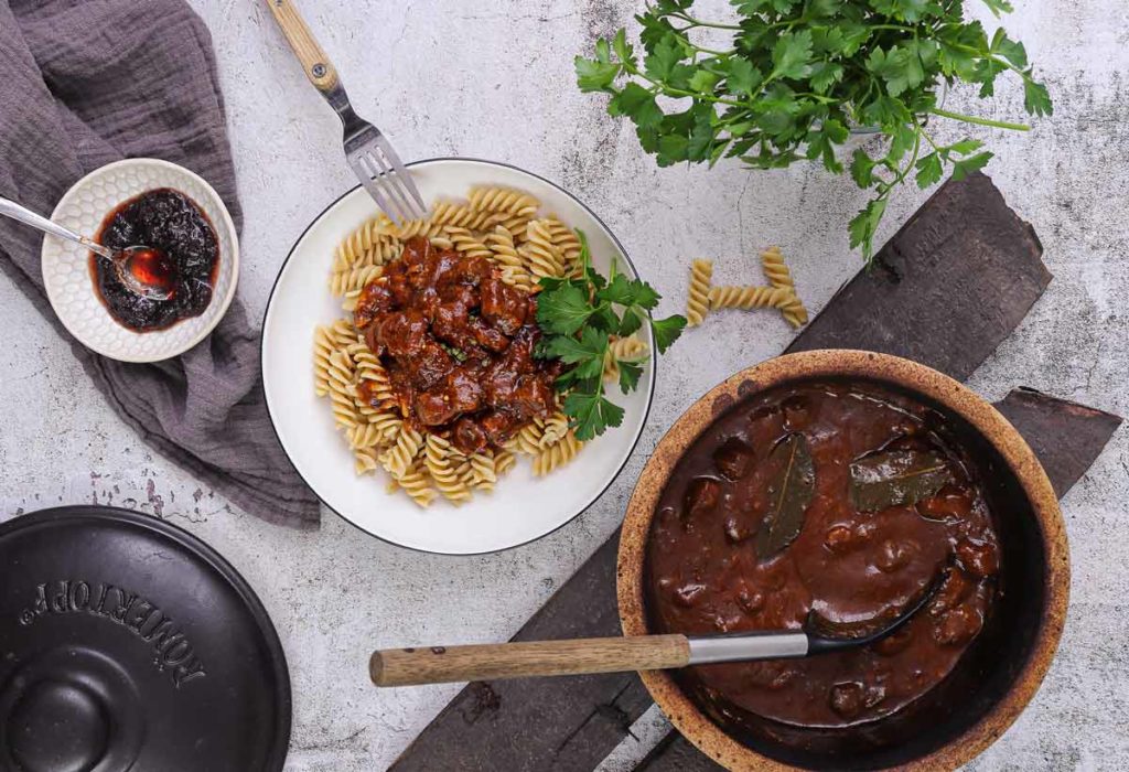 Teller mit Nudeln und Gulasch, daneben Topf mit Gulasch, Schüssel mit Soße, Petersilie, auf hellem Untergrund.