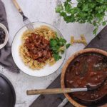 Teller mit Nudeln und Gulasch, daneben Topf mit Gulasch, Schüssel mit Soße, Petersilie, auf hellem Untergrund.