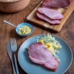 Schinkenstück auf Teller mit Kartoffelsalat, Gabel und Messer; Senfschale und Holzbrett mit Schinken im Hintergrund.