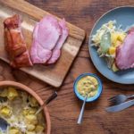 Tisch mit Kassler auf Holzbrett, Sauerkraut mit Kartoffeln in Schüssel, Senf, Teller mit Kassler und Beilagen.