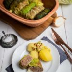 Teller mit Kohlrouladen, Kartoffeln und Gewürzen; dahinter eine Auflaufform mit weiteren Rouladen auf weißem Holztisch.