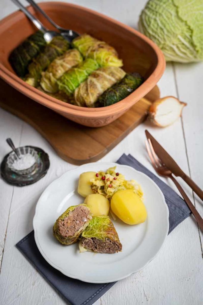 Teller mit Kohlrouladen, Kartoffeln und Gewürzen; dahinter eine Auflaufform mit weiteren Rouladen auf weißem Holztisch.