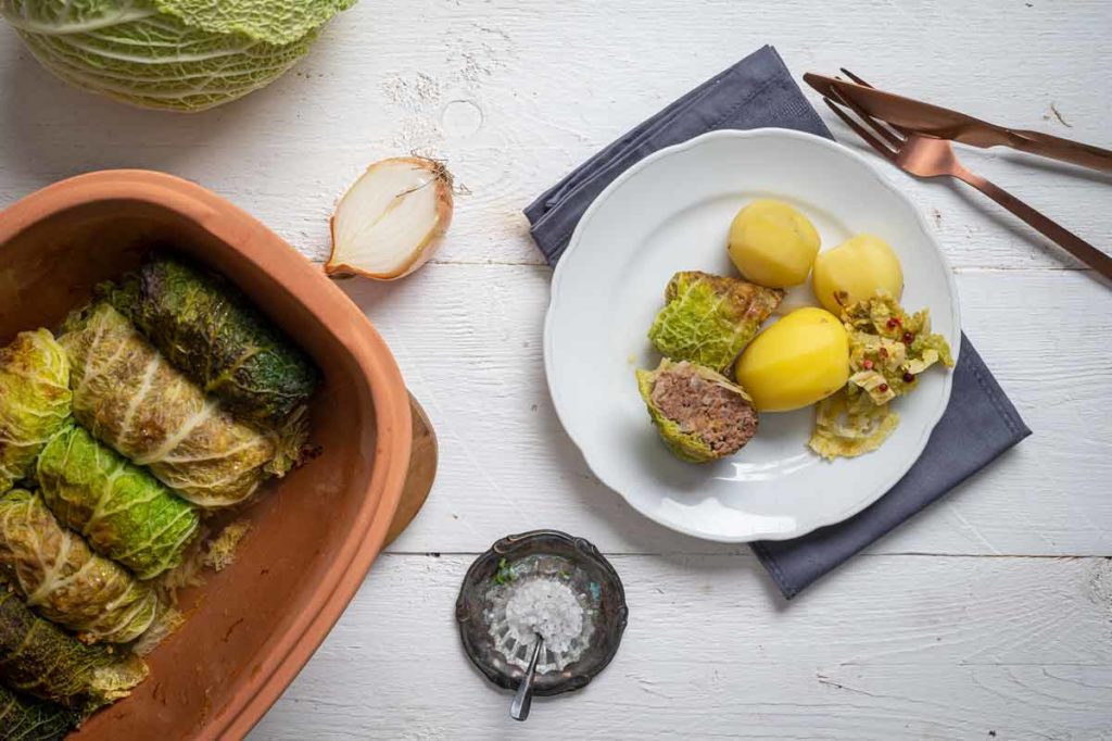 Gefüllte Wirsingrouladen mit Salzkartoffeln auf einem Teller, daneben ein Tontopf mit weiteren Rouladen und eine geschnittene Zwiebel.