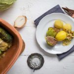 Gefüllte Wirsingrouladen mit Salzkartoffeln auf einem Teller, daneben ein Tontopf mit weiteren Rouladen und eine geschnittene Zwiebel.