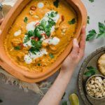 Hand hält eine Schale mit Currygericht, garniert mit Joghurt und Kräutern, daneben Naan, Cashews und Dip auf einem Tablett.