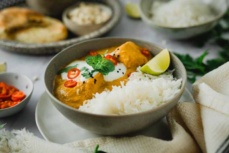 Schale mit Curry und Reis, garniert mit Chili, Joghurt und Minze, daneben Limettenscheibe und Brot im Hintergrund.