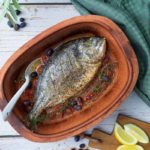 Gegrillter Fisch mit Tomaten und Oliven in Tonform, dekoriert mit Zitrone, Kräutern und einem grünen Tuch.