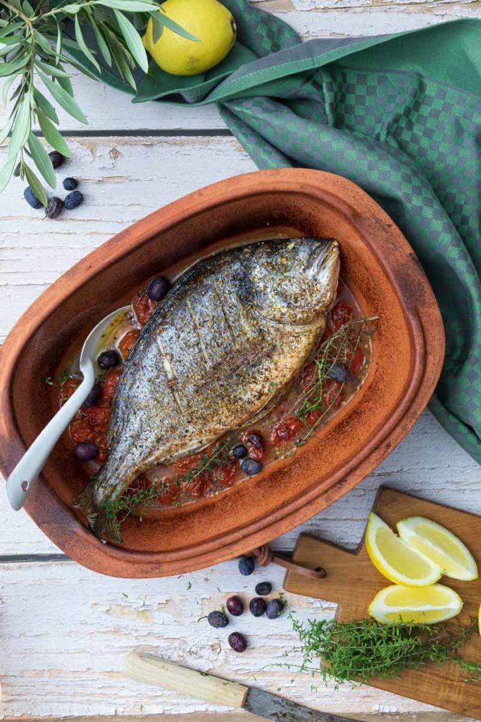 Gegrillter Fisch mit Tomaten und Oliven in Tonform, dekoriert mit Zitrone, Kräutern und einem grünen Tuch.